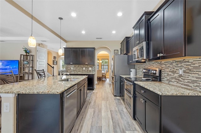 kitchen with appliances with stainless steel finishes, hanging light fixtures, light hardwood / wood-style floors, a kitchen island with sink, and sink