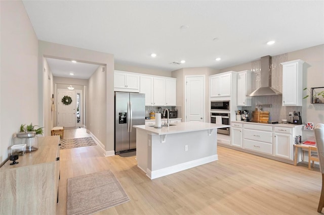 kitchen with light wood-type flooring, an island with sink, wall chimney range hood, and stainless steel appliances