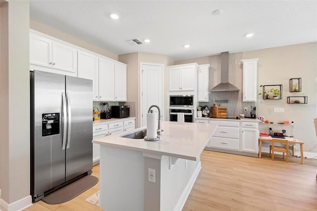 kitchen featuring appliances with stainless steel finishes, white cabinets, wall chimney exhaust hood, light wood-type flooring, and a center island with sink