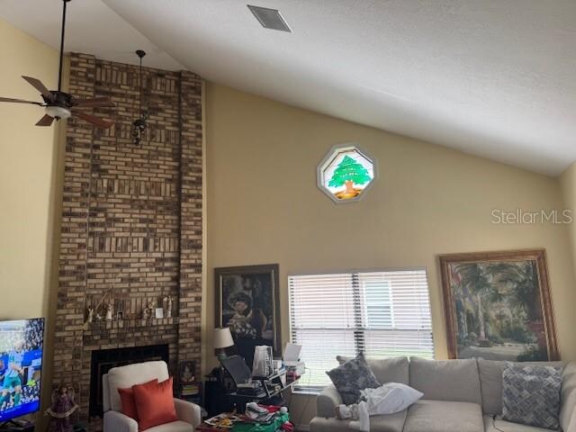 living room featuring a brick fireplace, ceiling fan, a textured ceiling, and vaulted ceiling