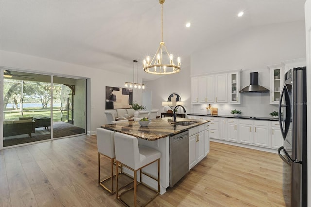 kitchen with wall chimney range hood, sink, white cabinetry, stainless steel appliances, and an island with sink