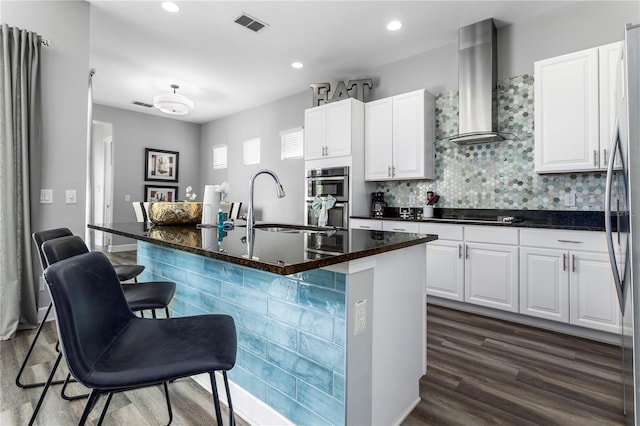 kitchen with wall chimney exhaust hood, a center island with sink, dark hardwood / wood-style floors, a kitchen bar, and white cabinetry