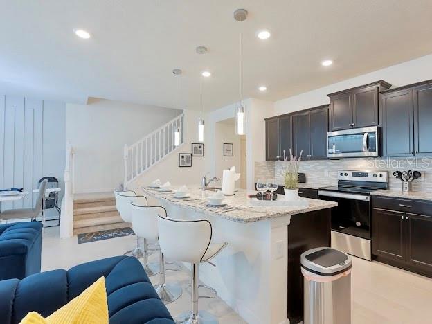 kitchen with an island with sink, light stone counters, stainless steel appliances, tasteful backsplash, and decorative light fixtures