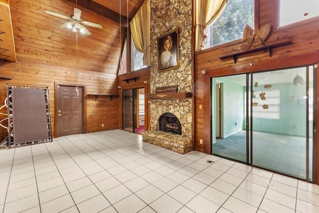 unfurnished living room with a stone fireplace, tile patterned flooring, ceiling fan, high vaulted ceiling, and wood walls