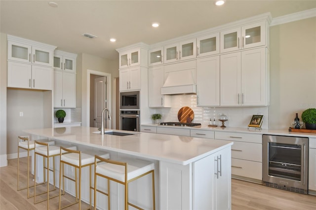 kitchen featuring a kitchen island with sink, stainless steel appliances, white cabinetry, beverage cooler, and premium range hood