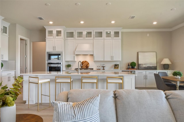 kitchen with an island with sink, stainless steel appliances, white cabinets, crown molding, and premium range hood