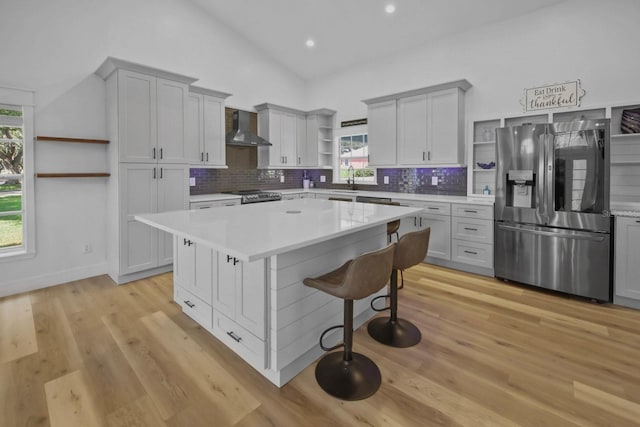 kitchen with a kitchen island, appliances with stainless steel finishes, a kitchen breakfast bar, wall chimney exhaust hood, and light hardwood / wood-style flooring