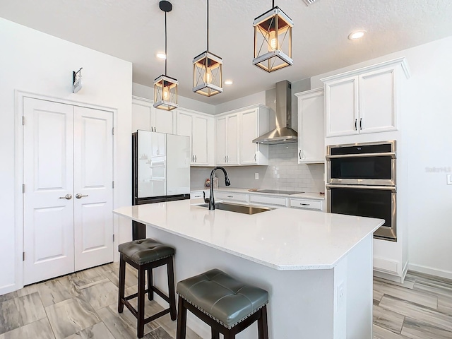 kitchen featuring a kitchen island with sink, sink, wall chimney exhaust hood, decorative light fixtures, and white cabinetry