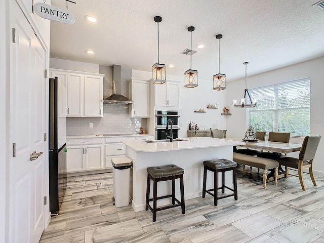 kitchen featuring wall chimney exhaust hood, a kitchen island with sink, pendant lighting, white cabinets, and a breakfast bar area