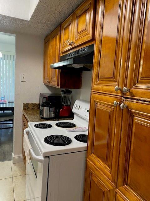 kitchen featuring electric stove, a textured ceiling, brown cabinetry, light tile patterned floors, and under cabinet range hood