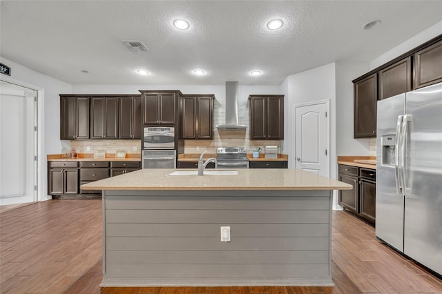 kitchen with wall chimney exhaust hood, appliances with stainless steel finishes, a textured ceiling, and a center island with sink