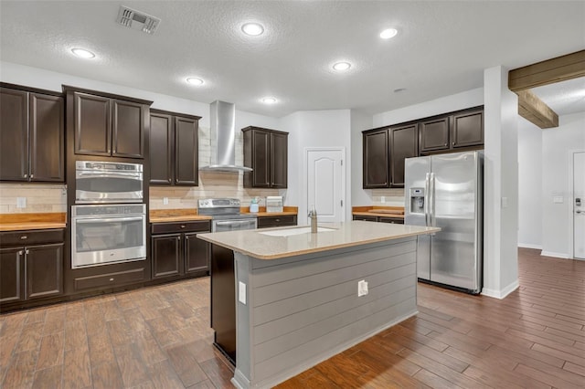 kitchen with wood-type flooring, wall chimney range hood, appliances with stainless steel finishes, a textured ceiling, and a center island with sink