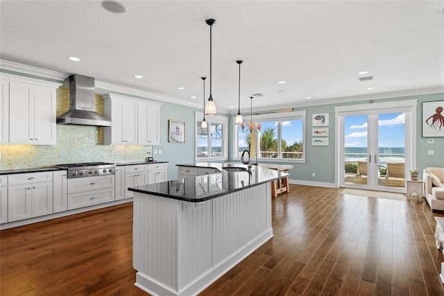 kitchen with wall chimney range hood, an island with sink, white cabinetry, stainless steel gas cooktop, and sink