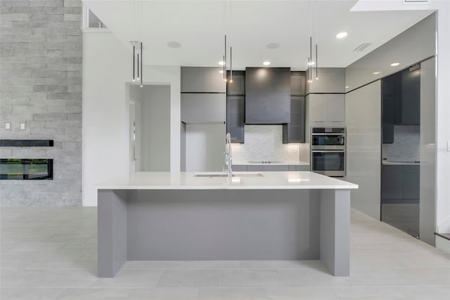 kitchen featuring a large island with sink, stainless steel double oven, hanging light fixtures, gray cabinetry, and backsplash