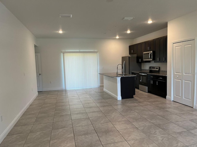 kitchen with appliances with stainless steel finishes, a kitchen island with sink, light stone counters, and light tile patterned floors