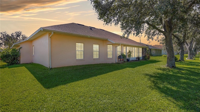 back of house at dusk featuring a shingled roof, a yard, and stucco siding