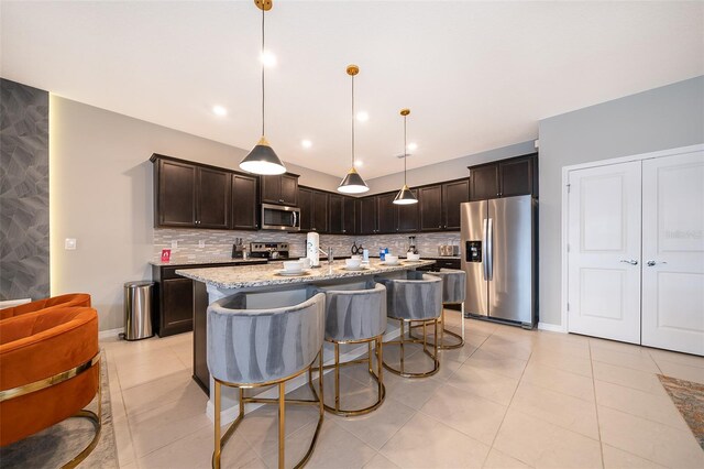 kitchen featuring light stone countertops, an island with sink, backsplash, stainless steel appliances, and pendant lighting