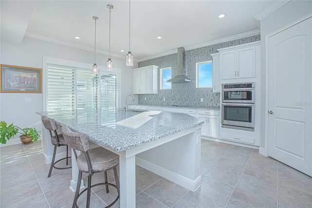 kitchen with wall chimney range hood, hanging light fixtures, white cabinetry, a kitchen island with sink, and stainless steel double oven