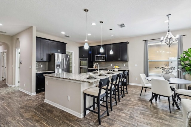 kitchen featuring hanging light fixtures, backsplash, appliances with stainless steel finishes, a kitchen island with sink, and dark wood-type flooring