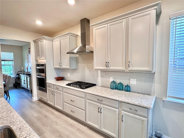 kitchen with gas stovetop, stainless steel double oven, wall chimney exhaust hood, light wood-type flooring, and light stone counters