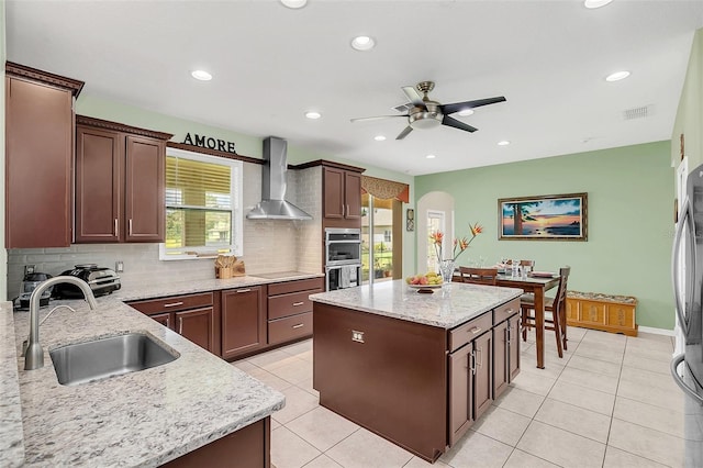 kitchen featuring wall chimney range hood, light stone counters, a kitchen island, sink, and stainless steel double oven