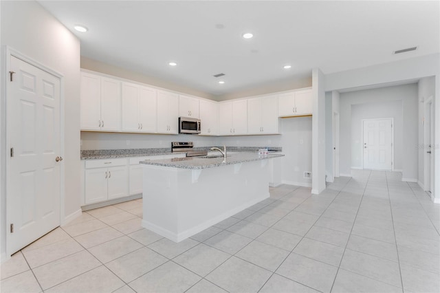 kitchen with light stone countertops, stainless steel appliances, white cabinets, and an island with sink