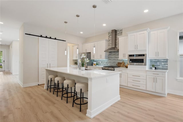 kitchen featuring an island with sink, stainless steel appliances, white cabinets, a barn door, and wall chimney range hood