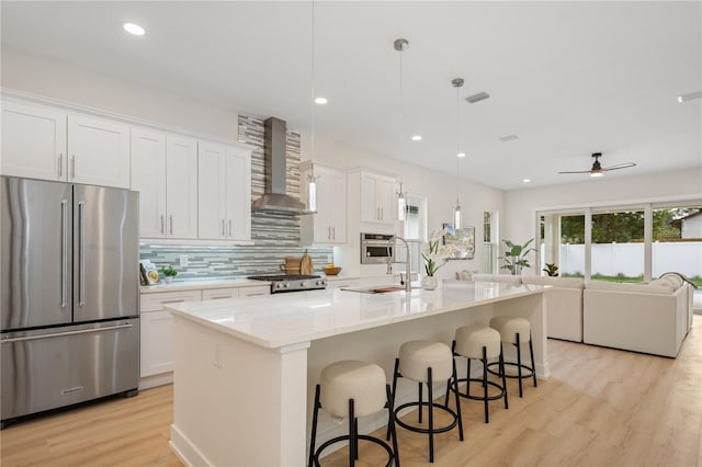 kitchen featuring a kitchen island with sink, wall chimney range hood, white cabinetry, appliances with stainless steel finishes, and light wood finished floors