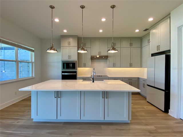 kitchen with an island with sink, stainless steel appliances, sink, light stone countertops, and light wood-type flooring