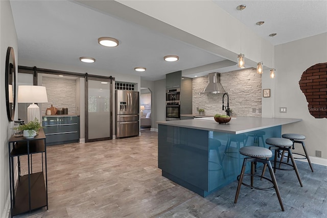 kitchen featuring wall chimney exhaust hood, kitchen peninsula, a barn door, stainless steel fridge with ice dispenser, and pendant lighting