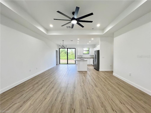 unfurnished living room featuring a tray ceiling, sink, light wood-type flooring, and ceiling fan with notable chandelier