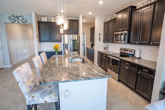 kitchen featuring light tile patterned floors, hanging light fixtures, appliances with stainless steel finishes, a kitchen island with sink, and dark brown cabinetry