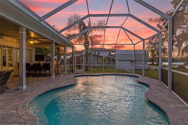 pool at dusk with a yard, a patio, glass enclosure, and ceiling fan