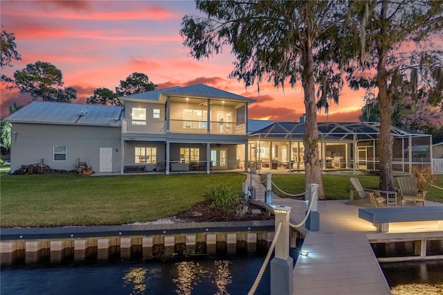 back house at dusk with a balcony, a water view, a lawn, and glass enclosure