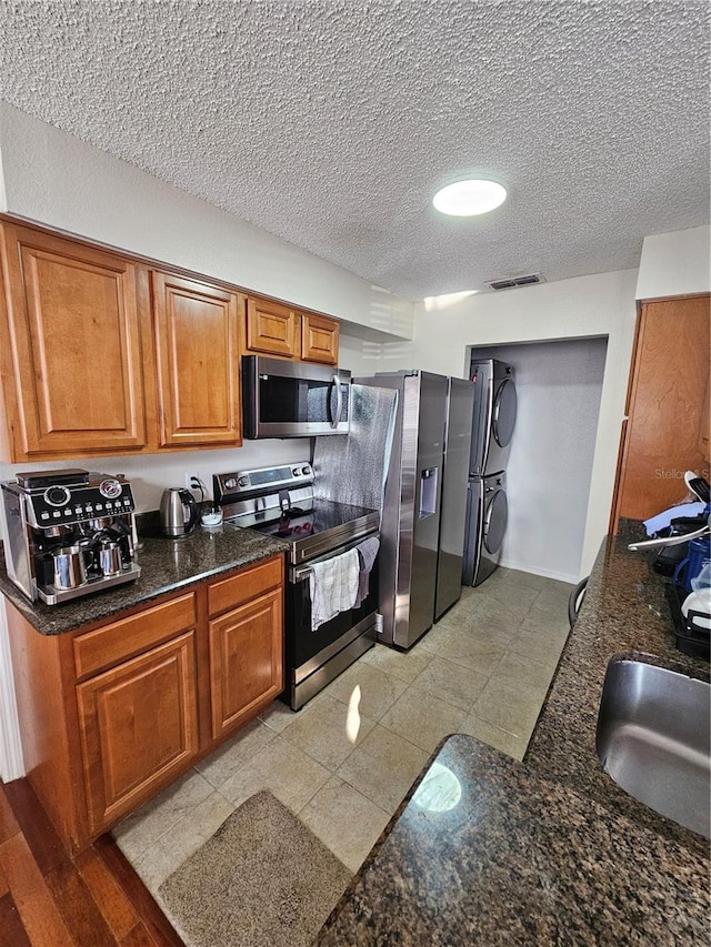 kitchen with appliances with stainless steel finishes, dark stone counters, a textured ceiling, and stacked washing maching and dryer