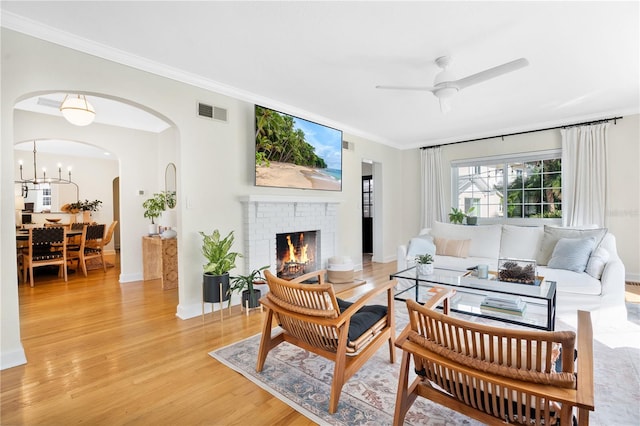 living room with a brick fireplace, ceiling fan with notable chandelier, crown molding, and light hardwood / wood-style flooring