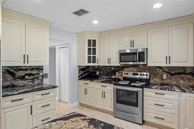 kitchen with cream cabinets, light tile patterned floors, appliances with stainless steel finishes, backsplash, and dark stone countertops