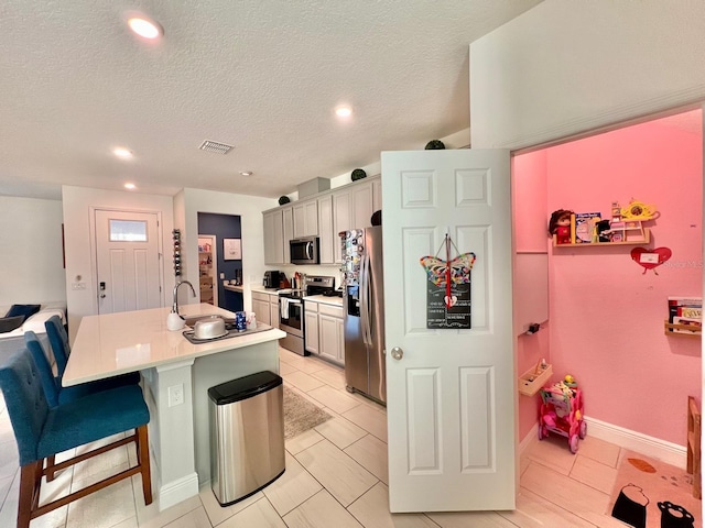 kitchen with a kitchen island with sink, stainless steel appliances, a kitchen bar, gray cabinets, and a textured ceiling