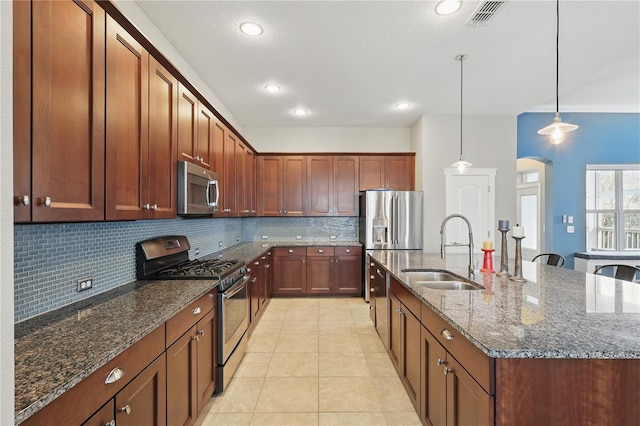 kitchen featuring a center island with sink, dark stone counters, appliances with stainless steel finishes, hanging light fixtures, and a sink