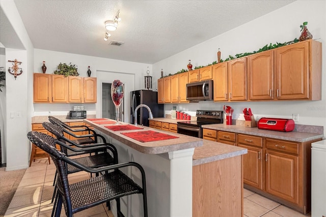 kitchen with light tile patterned flooring, appliances with stainless steel finishes, a kitchen breakfast bar, a textured ceiling, and a kitchen island with sink