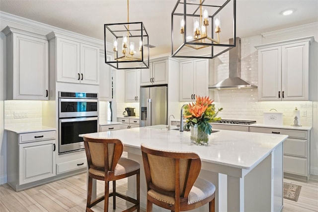 kitchen featuring appliances with stainless steel finishes, wall chimney range hood, pendant lighting, light hardwood / wood-style flooring, and an island with sink