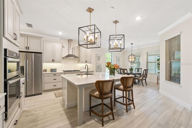 kitchen with stainless steel appliances, sink, wall chimney range hood, decorative light fixtures, and a center island with sink