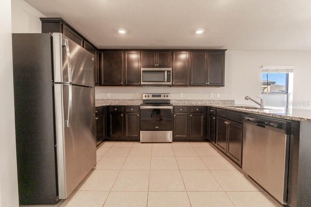 kitchen featuring dark brown cabinets, light tile patterned floors, appliances with stainless steel finishes, light stone countertops, and sink