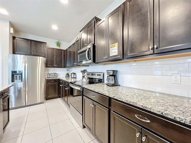 kitchen with dark brown cabinets, backsplash, light tile patterned floors, light stone countertops, and stainless steel appliances