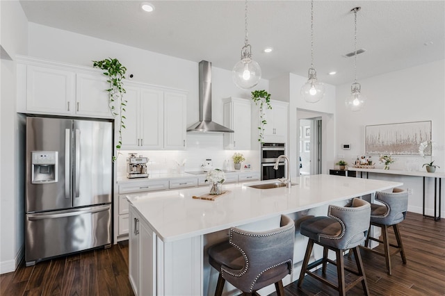 kitchen with an island with sink, stainless steel appliances, wall chimney exhaust hood, pendant lighting, and white cabinets