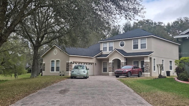 traditional-style house with a garage, stone siding, decorative driveway, stucco siding, and a front yard