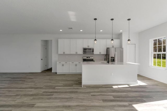 kitchen with light wood-type flooring, stainless steel appliances, decorative light fixtures, white cabinets, and a kitchen island with sink