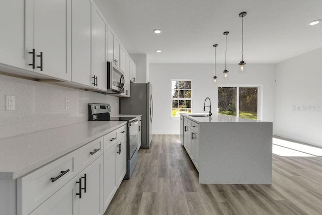 kitchen with appliances with stainless steel finishes, hanging light fixtures, light hardwood / wood-style floors, white cabinets, and a kitchen island with sink