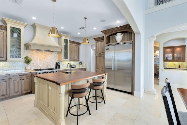 kitchen with custom exhaust hood, stainless steel built in fridge, a center island with sink, hanging light fixtures, and wood counters