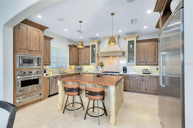 kitchen with an island with sink, custom range hood, built in appliances, pendant lighting, and sink